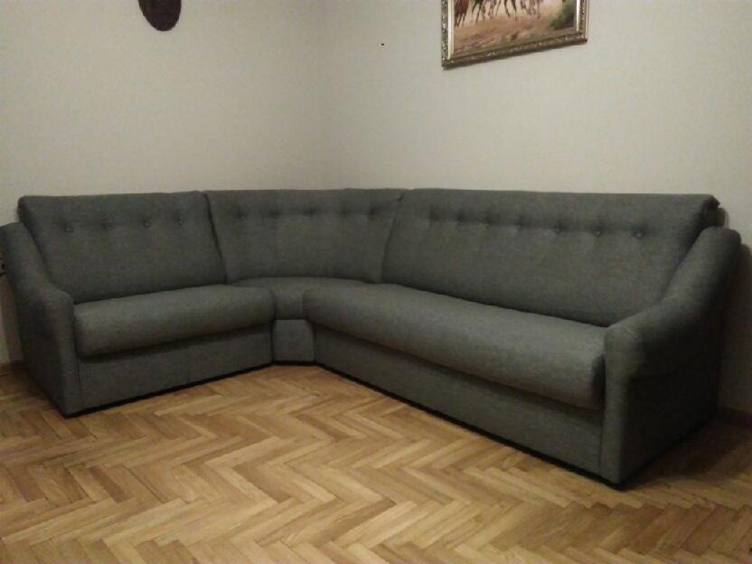 Szary narożnik tapicerowany tkaniną z guzikami na oparciu, ustawiony na parkiecie jodełkowym, z obrazem koni na ścianie w tle.