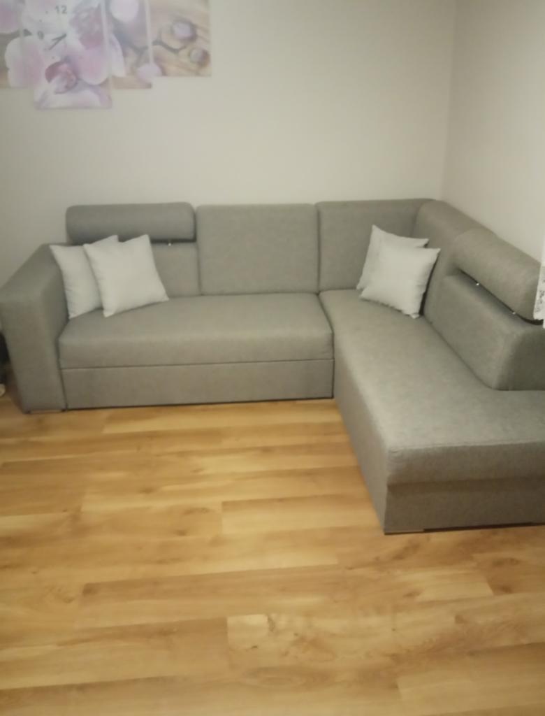 Szary narożnik z regulowanymi zagłówkami i białymi poduszkami na drewnianej podłodze, sfotografowany w rogu pokoju pod kątem.