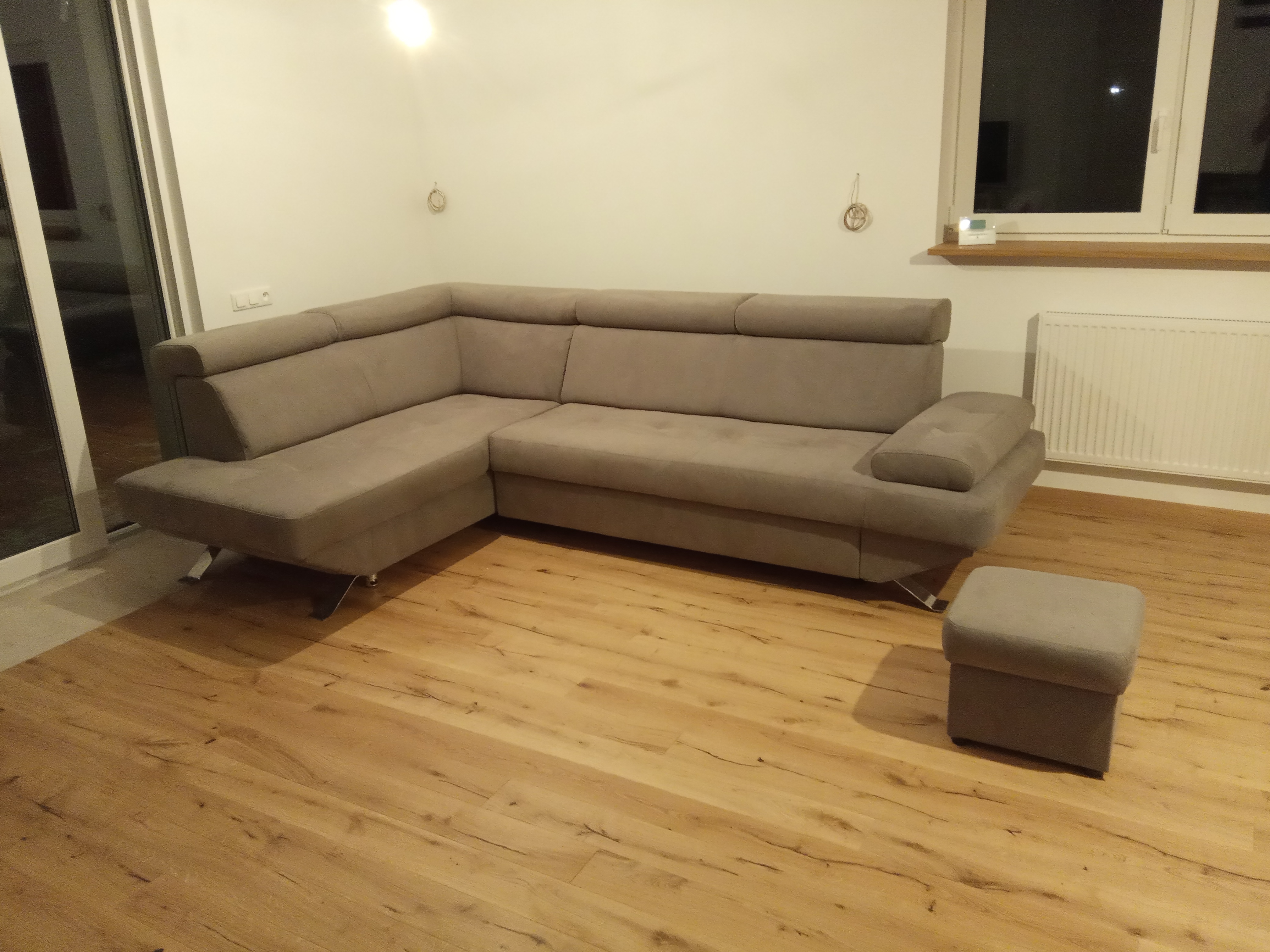 Beżowy, narożny zestaw wypoczynkowy z pufą na drewnianej podłodze w jasnym wnętrzu.