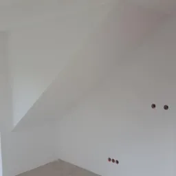 Białe, świeżo pomalowane wnętrze poddasza ze skosami, widoczne puszki elektryczne bez osprzętu i fragment okna z widokiem na zewnątrz.