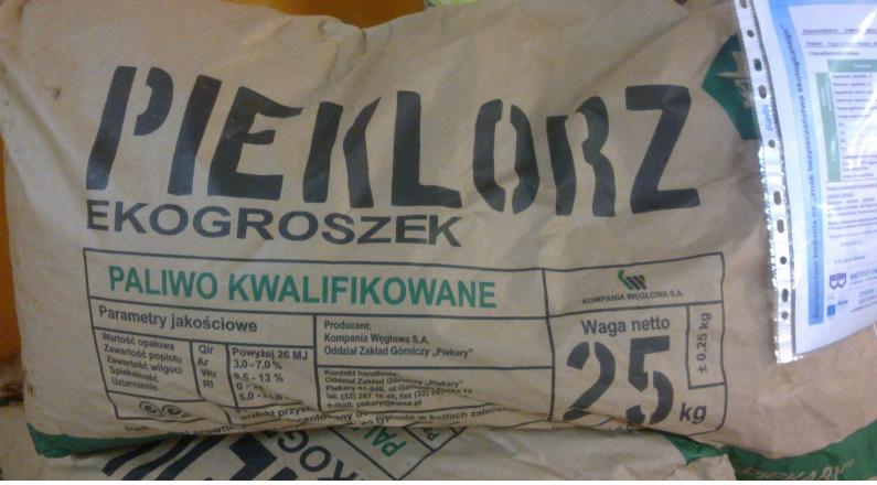 Worek z ekogroszkiem marki PIEKLORZ, z widocznymi parametrami jakościowymi i wagą netto 25 kg, na drugim planie częściowo widoczny inny worek oraz dokument w koszulce.
