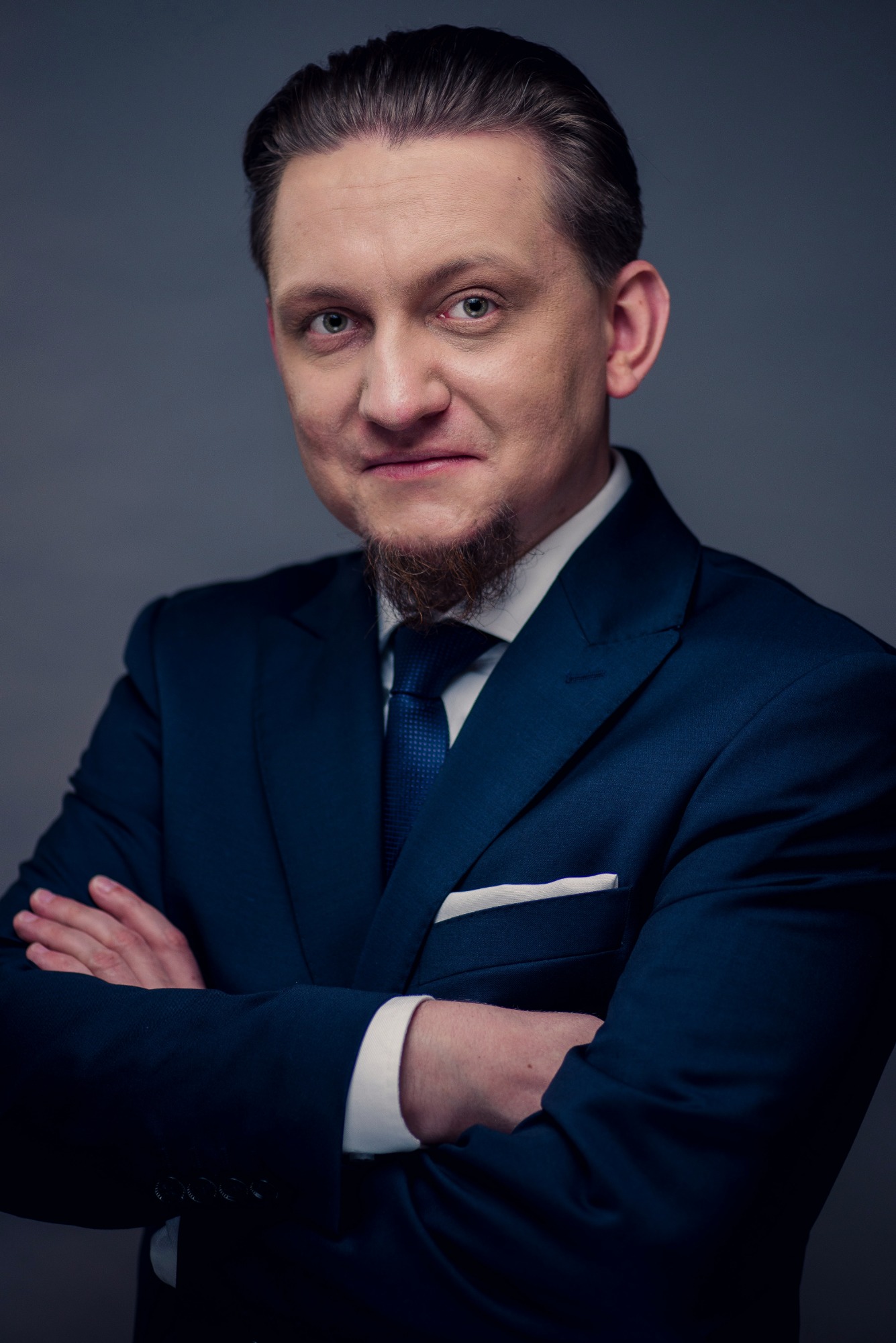 Portret mężczyzny w granatowym garniturze, z rękami skrzyżowanymi na piersiach, patrzącego w obiektyw na tle szarego tła.