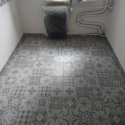 Ułożona podłoga z płytek ceramicznych w odcieniach szarości z wzorem patchworkowym, grzejnik zabezpieczony folią malarską, białe rury grzewcze na ścianie.