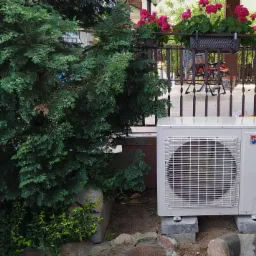 Zewnętrzna jednostka pompy ciepła marki Therm, umieszczona przy ścianie domu obok krzewu iglastego i kwiatów w donicy na balkonie.