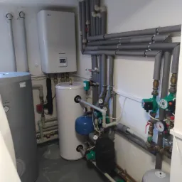 Kotłownia z dwoma zbiornikami akumulacyjnymi, kotłem gazowym i rurami izolowanymi termicznie, widoczne manometry i pompy obiegowe.