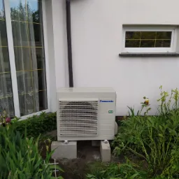 Zewnętrzna jednostka klimatyzacji Panasonic R32 zamontowana na betonowych bloczkach przy białej ścianie domu, otoczona zielenią i kwiatami, widok z poziomu gruntu.