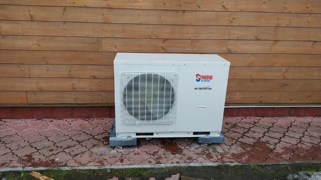 Zewnętrzna jednostka pompy ciepła marki STHERM DC Inverter, umieszczona na betonowych podstawach przed drewnianą elewacją budynku, na wilgotnej kostce brukowej.