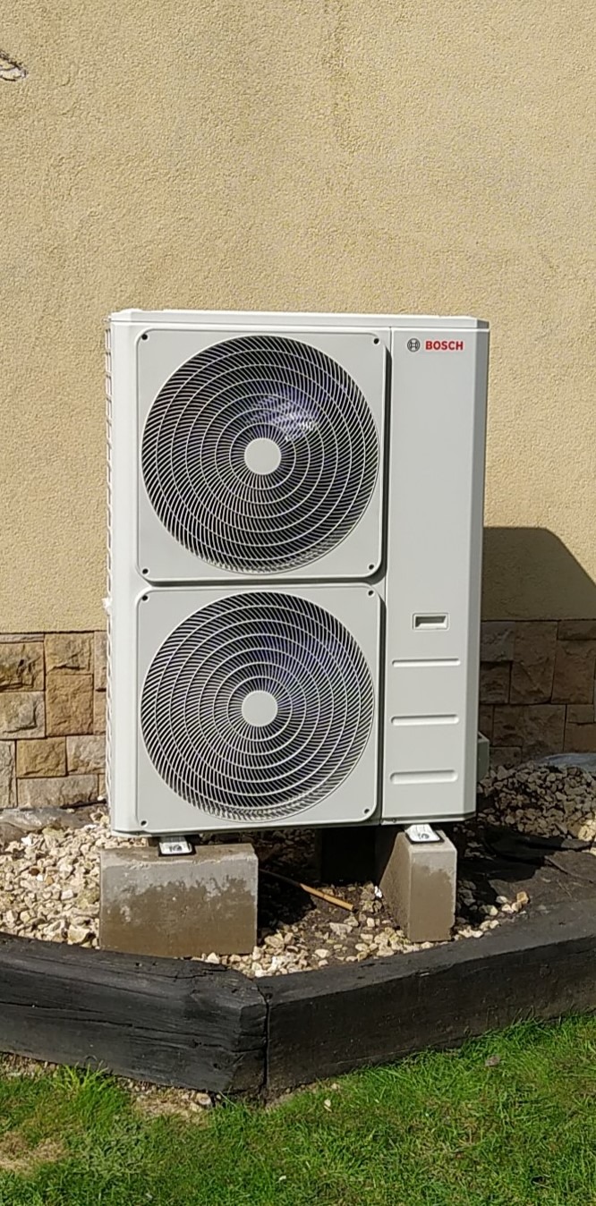 Biała, dwuczęściowa pompa ciepła marki Bosch, zamontowana na betonowych bloczkach przed budynkiem o jasnej elewacji i kamiennej podmurówce.