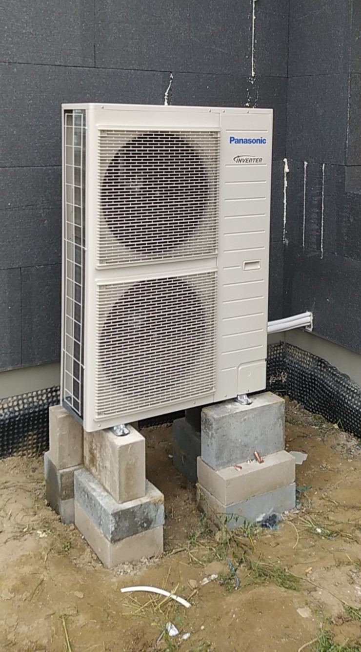 Zewnętrzna jednostka klimatyzacji Panasonic Inverter zamontowana na prowizorycznej podstawie z betonowych bloczków przy ścianie budynku z szarym ociepleniem.