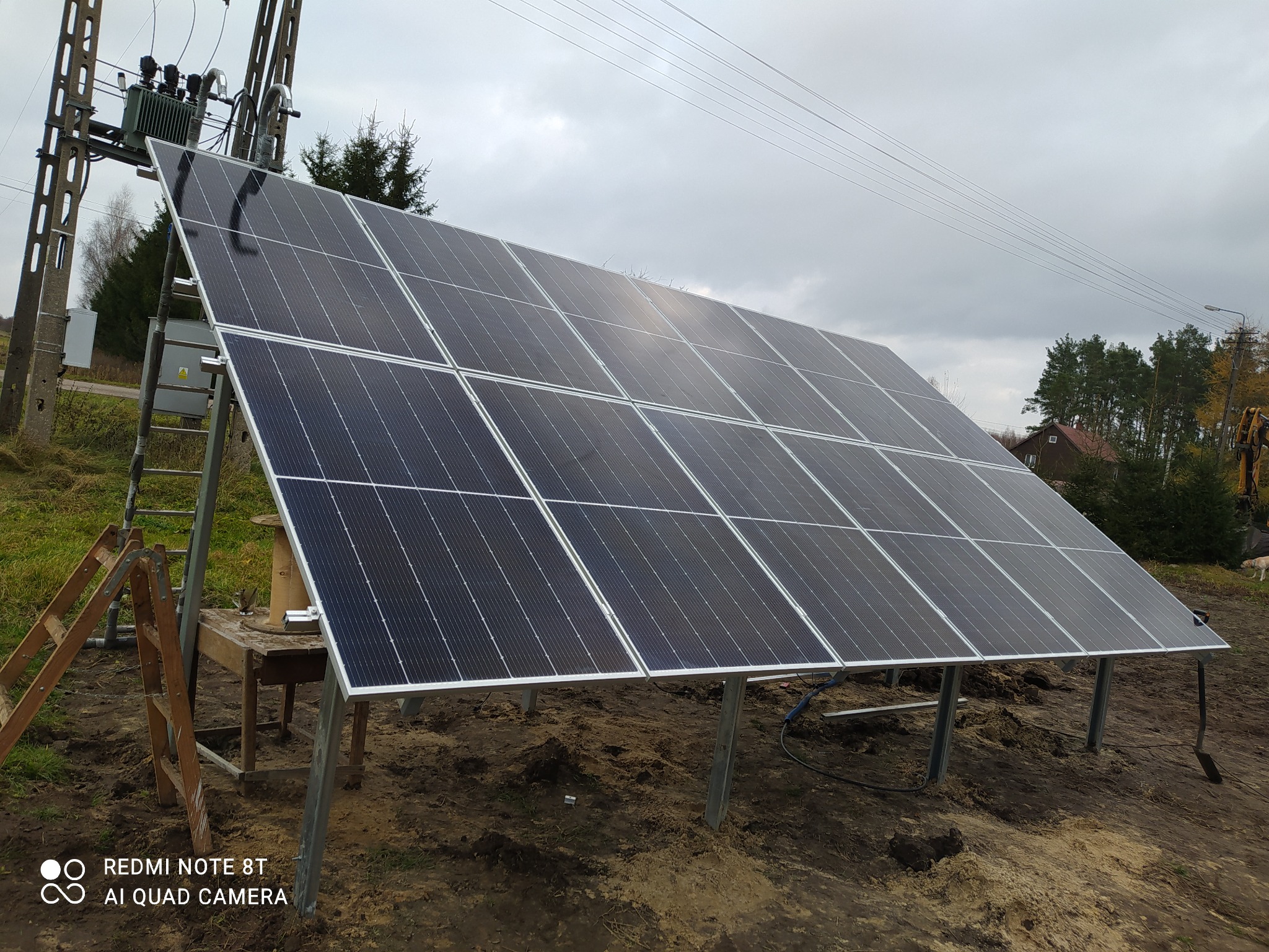 Panel słoneczny na metalowej konstrukcji, ustawiony na gruncie, obok drabina i stół warsztatowy, w tle słup energetyczny i pochmurne niebo.