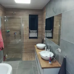 Nowoczesna łazienka z dwoma umywalkami nablatowymi, kabiną prysznicową typu walk-in, drewnianym blatem i szarą glazurą na ścianach.