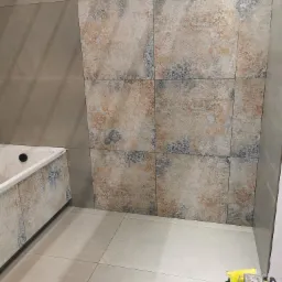 Wykończona łazienka z beżowo-złotymi płytkami dekoracyjnymi na ścianie i szarymi płytkami podłogowymi. Widoczny fragment wanny i akcesoria glazurnicze.