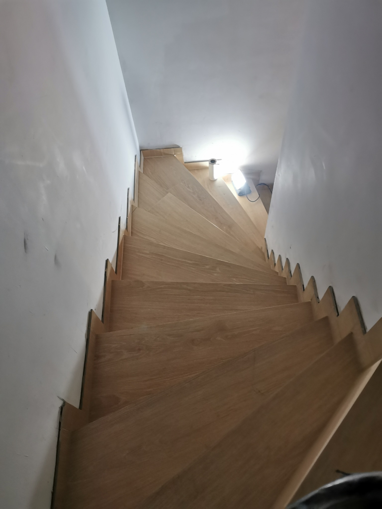 Jasne, drewniane schody z widocznymi stopniami, zamontowane w białym korytarzu, oświetlone lampą budowlaną. Ujęcie z góry.