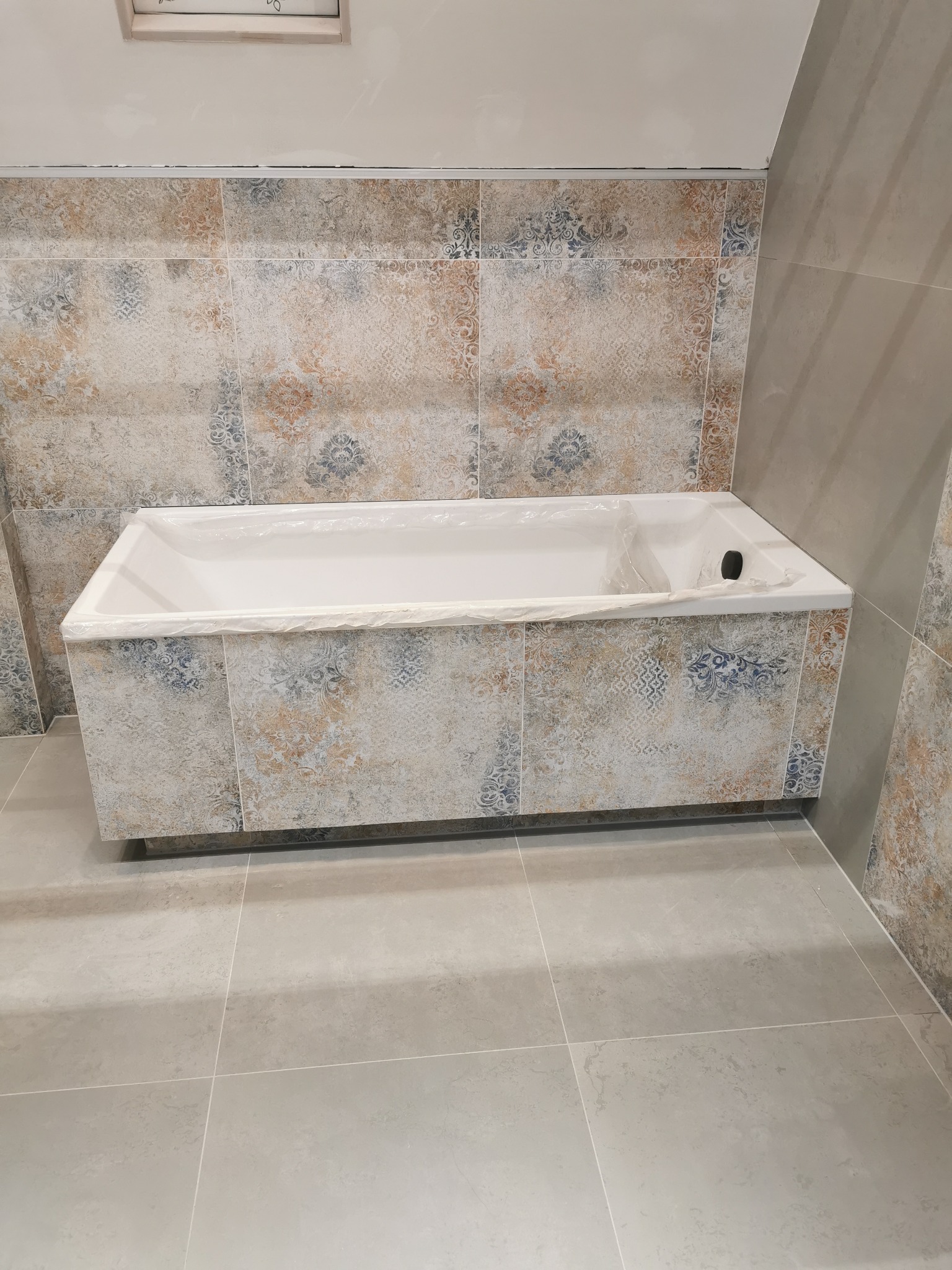 Wykończona łazienka z wanną obudowaną płytkami w mozaikę i jasnoszarą podłogą. Wanna zabezpieczona folią ochronną. Widoczne detale wykończenia.