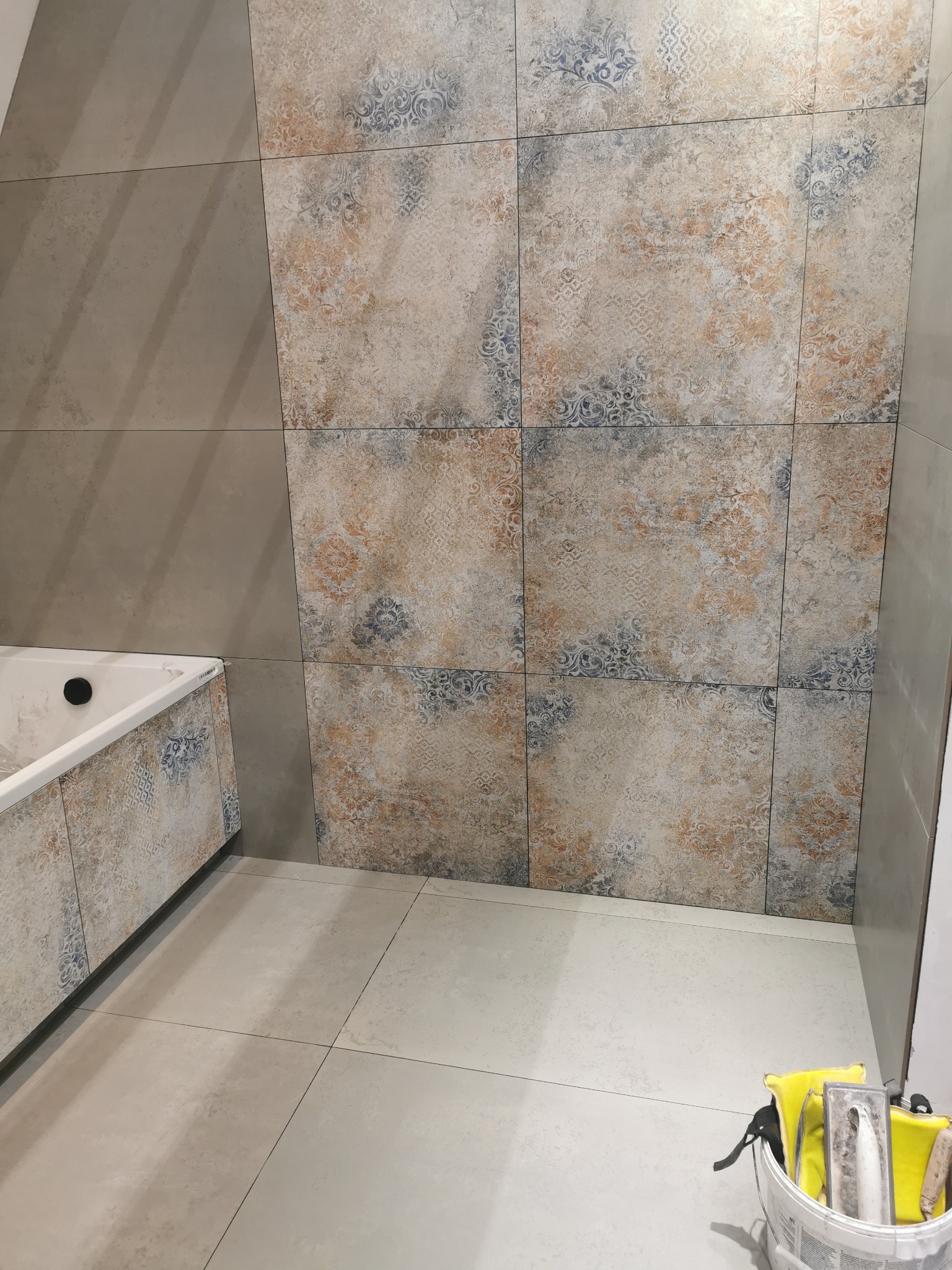 Wykończona łazienka z beżowo-złotymi płytkami dekoracyjnymi na ścianie i szarymi płytkami podłogowymi. Widoczny fragment wanny i akcesoria glazurnicze.