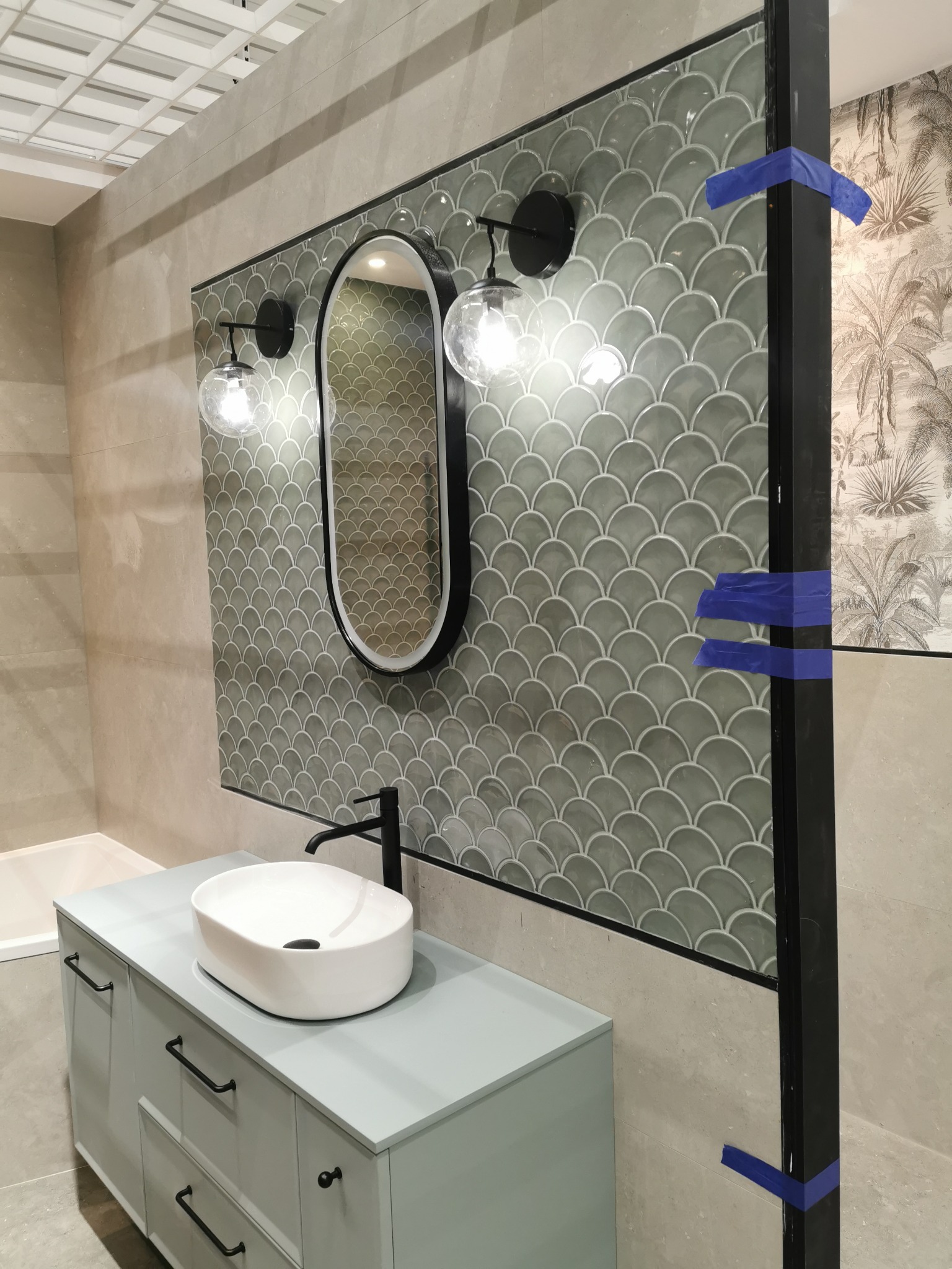 Wykończona łazienka z umywalką nablatową, lustrem w czarnej ramie i płytkami w kształcie rybiej łuski. Fragment parawanu nawannowego zabezpieczony taśmą.