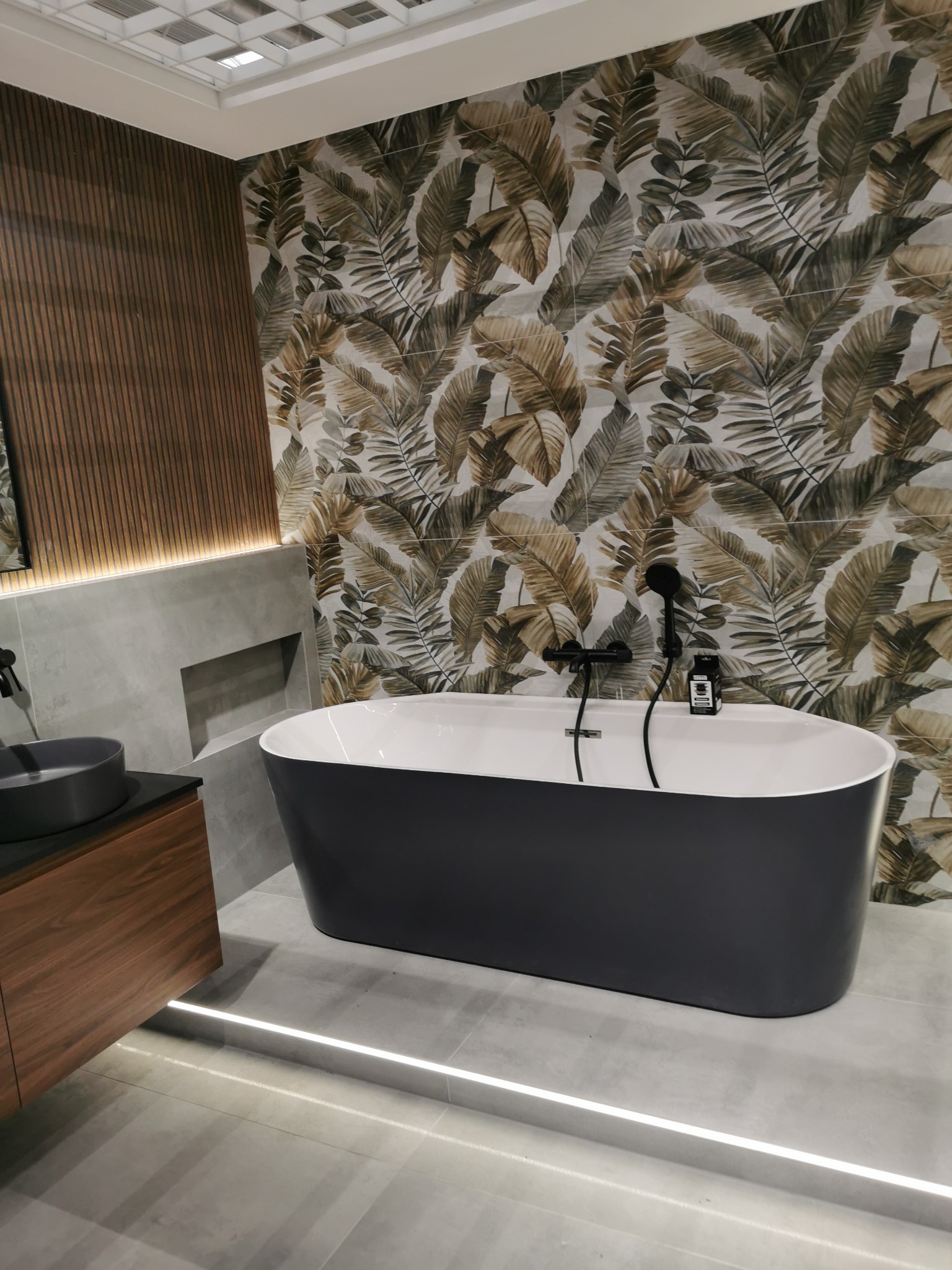 Nowoczesna łazienka z wolnostojącą wanną i płytkami w liście. Podświetlenie LED podkreśla gładkie, szare płytki podłogowe. Drewniane akcenty dodają ciepła.