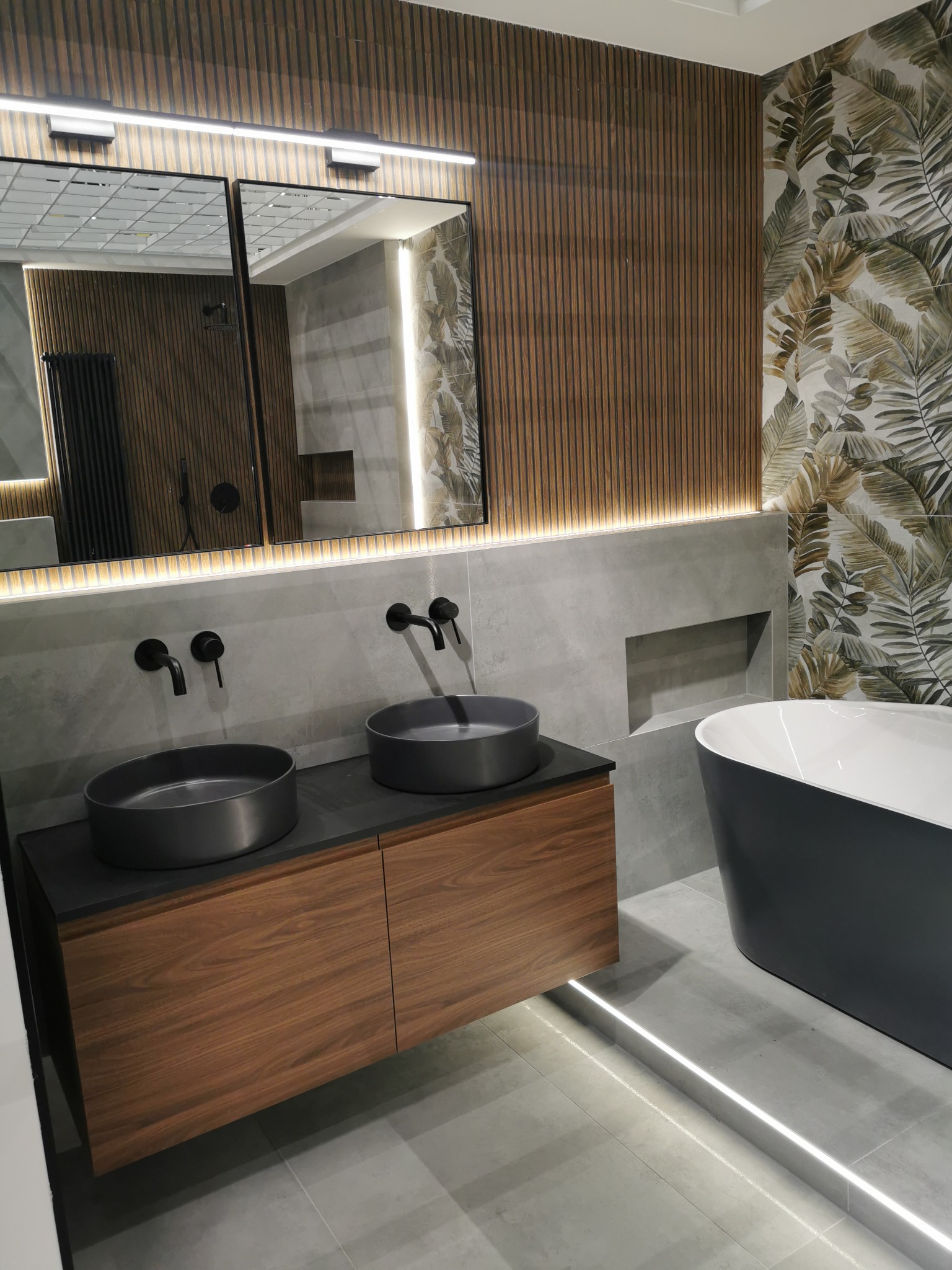 Nowoczesna łazienka z drewnianą szafką pod umywalki, szarą wanną i akcentem w postaci tapety w liście. Oświetlenie LED podkreśla minimalistyczny design.