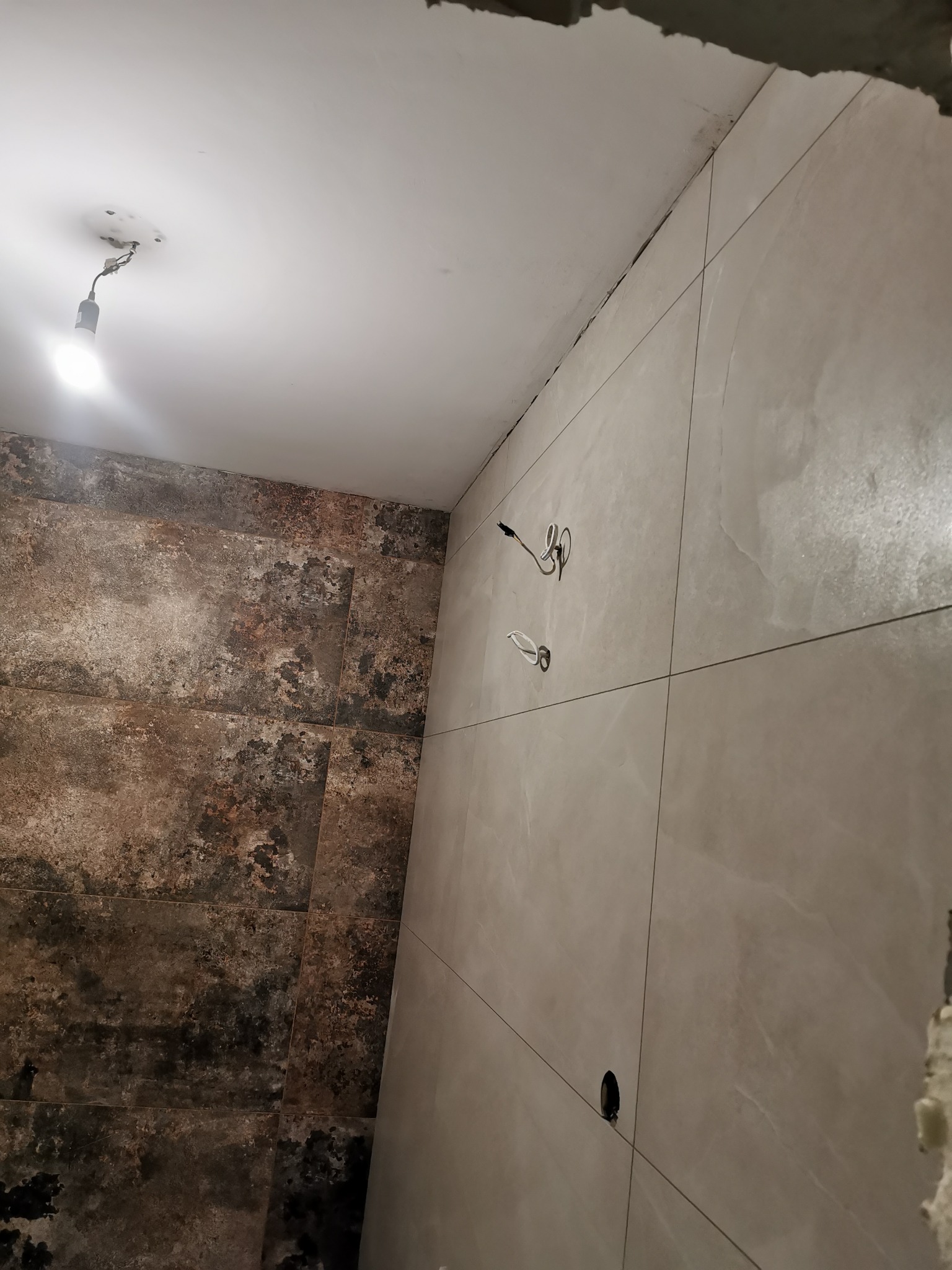 Układanie płytek w łazience: fragment ściany z beżowych płytek i brązowych płytek imitujących rdzę, widoczne kable elektryczne i sufit z żarówką.