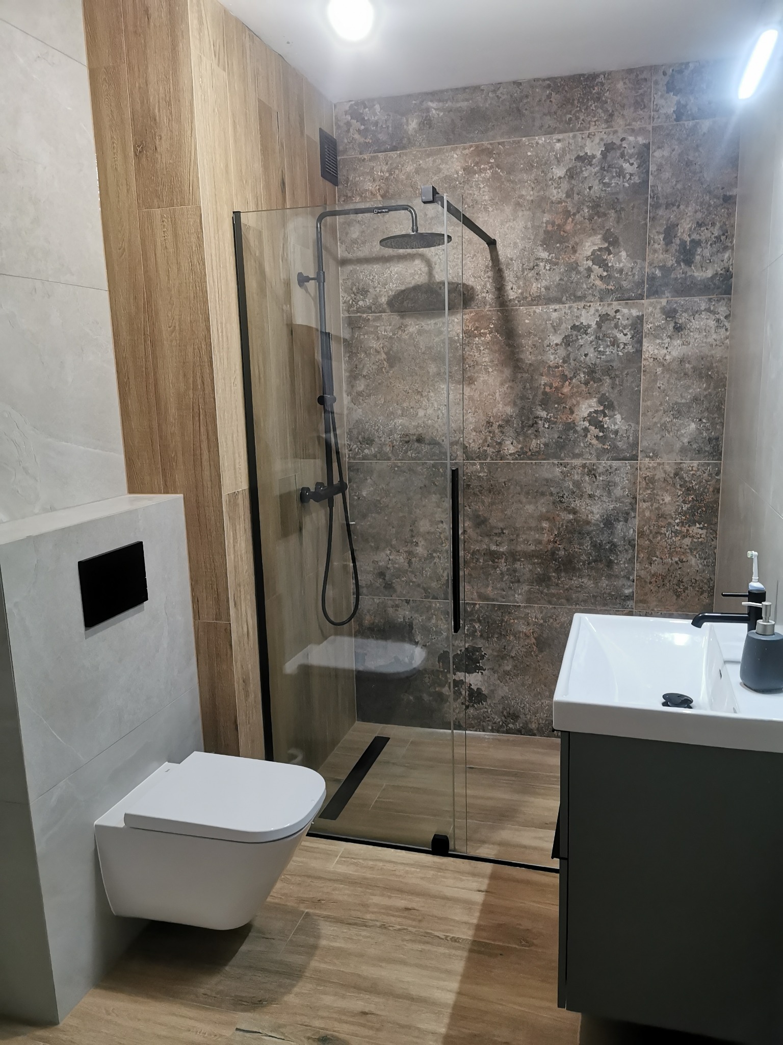 Nowoczesna łazienka po remoncie: kabina prysznicowa typu walk-in z czarną armaturą, umywalka z szafką i podwieszana toaleta. Płytki imitujące drewno i kamień.
