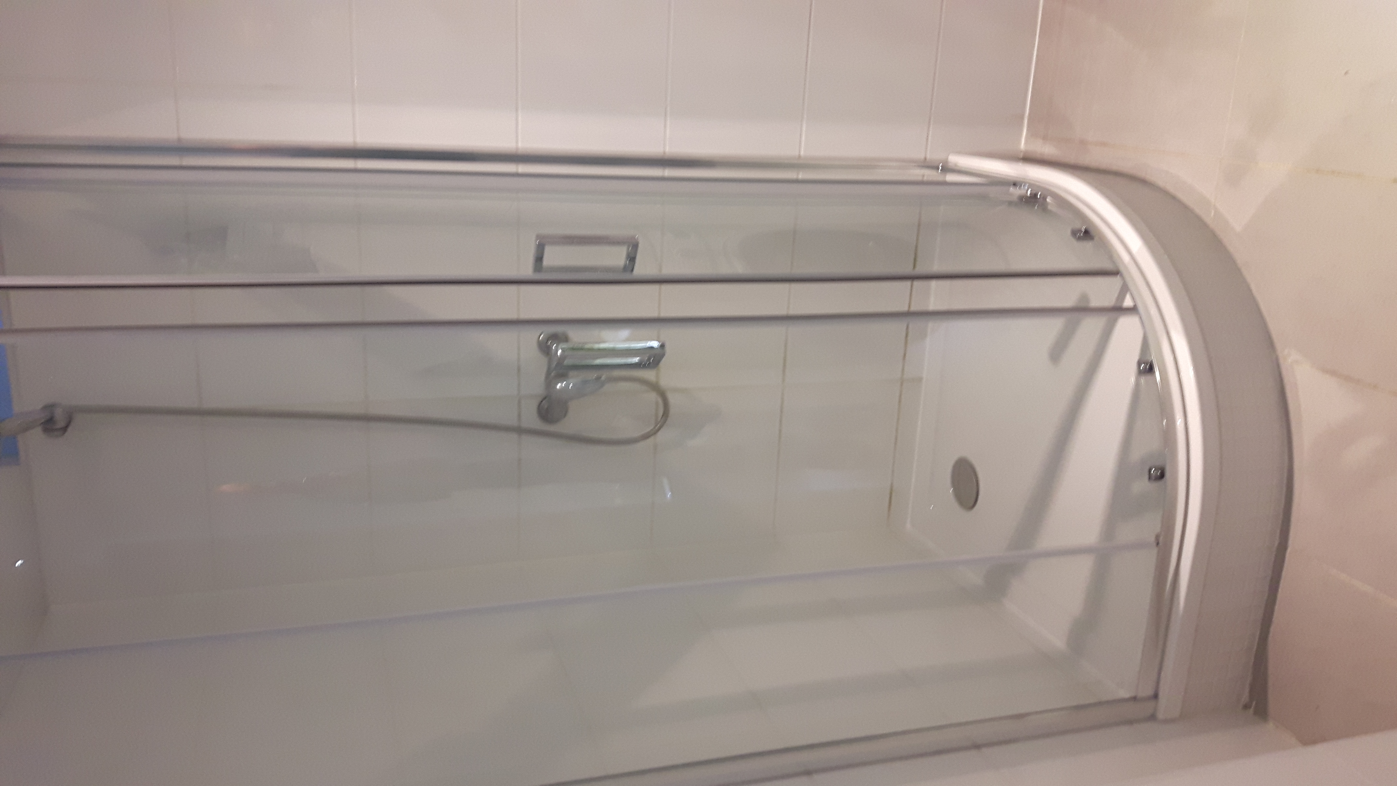 Szklana kabina prysznicowa z przesuwnymi drzwiami, widoczna bateria i wąż prysznicowy, białe płytki na ścianach, wnętrze kabiny z odpływem.