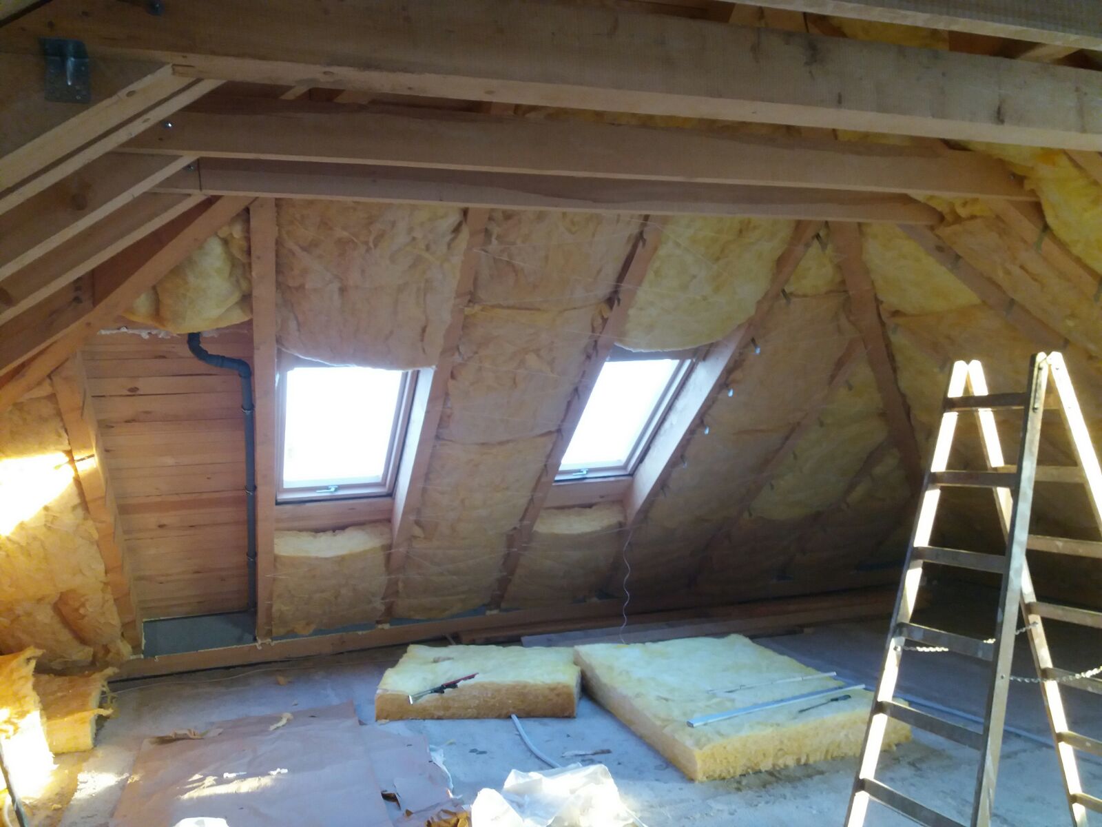 Poddasze w trakcie ocieplania wełną mineralną, widoczne okna dachowe, drewniana konstrukcja więźby dachowej oraz drabina oparta o ścianę.