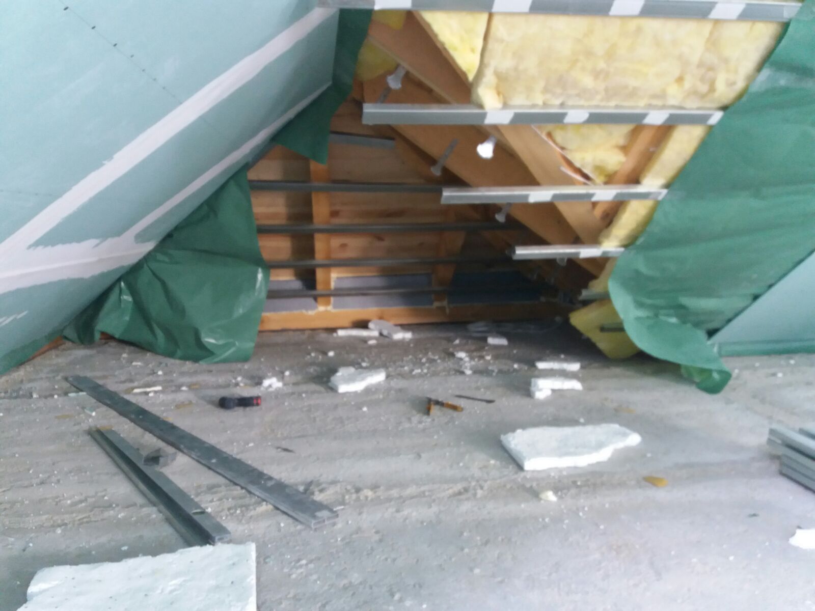 Poddasze w trakcie izolacji: widoczna wełna mineralna między krokwiami, częściowo zakryta folią ochronną i płytą gipsowo-kartonową. Na podłodze leżą fragmenty materiału izolacyjnego i narzędzia.