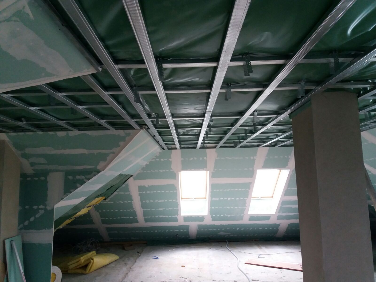 Wnętrze poddasza w trakcie prac wykończeniowych: widoczne metalowe profile sufitowe, częściowo zamontowane płyty gipsowo-kartonowe na ścianach ze szpachlowanymi łączeniami oraz okna dachowe.