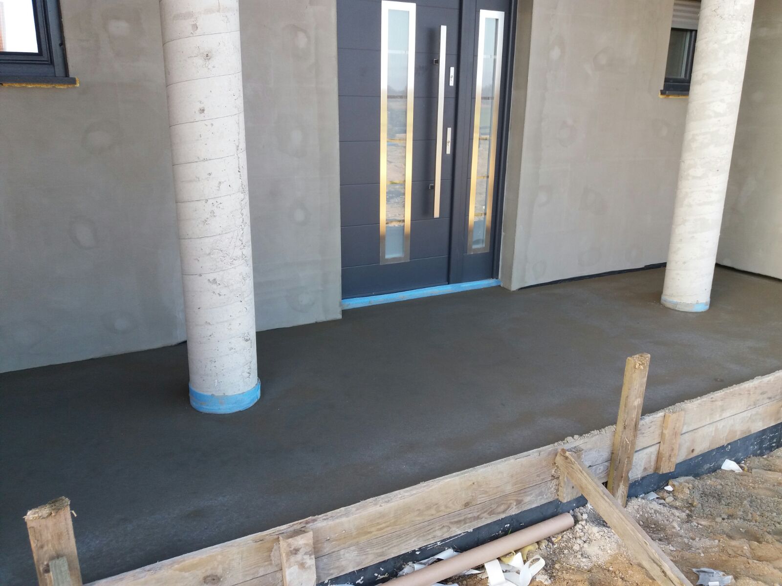 Świeżo wylana, szara posadzka przed wejściem do domu z nowoczesnymi drzwiami, zabezpieczona niebieską folią przy filarach i drzwiach, z widocznym drewnianym szalunkiem i elementami izolacji.