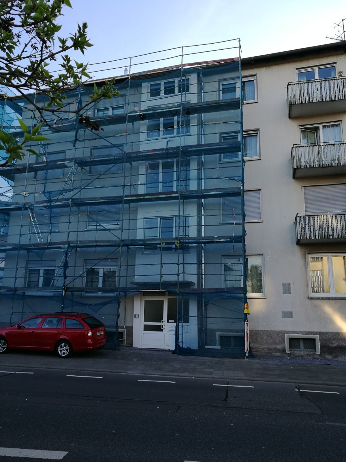 Budynek mieszkalny w trakcie prac elewacyjnych, osłonięty siatką rusztowania, z widocznym czerwonym samochodem osobowym zaparkowanym przed budynkiem.