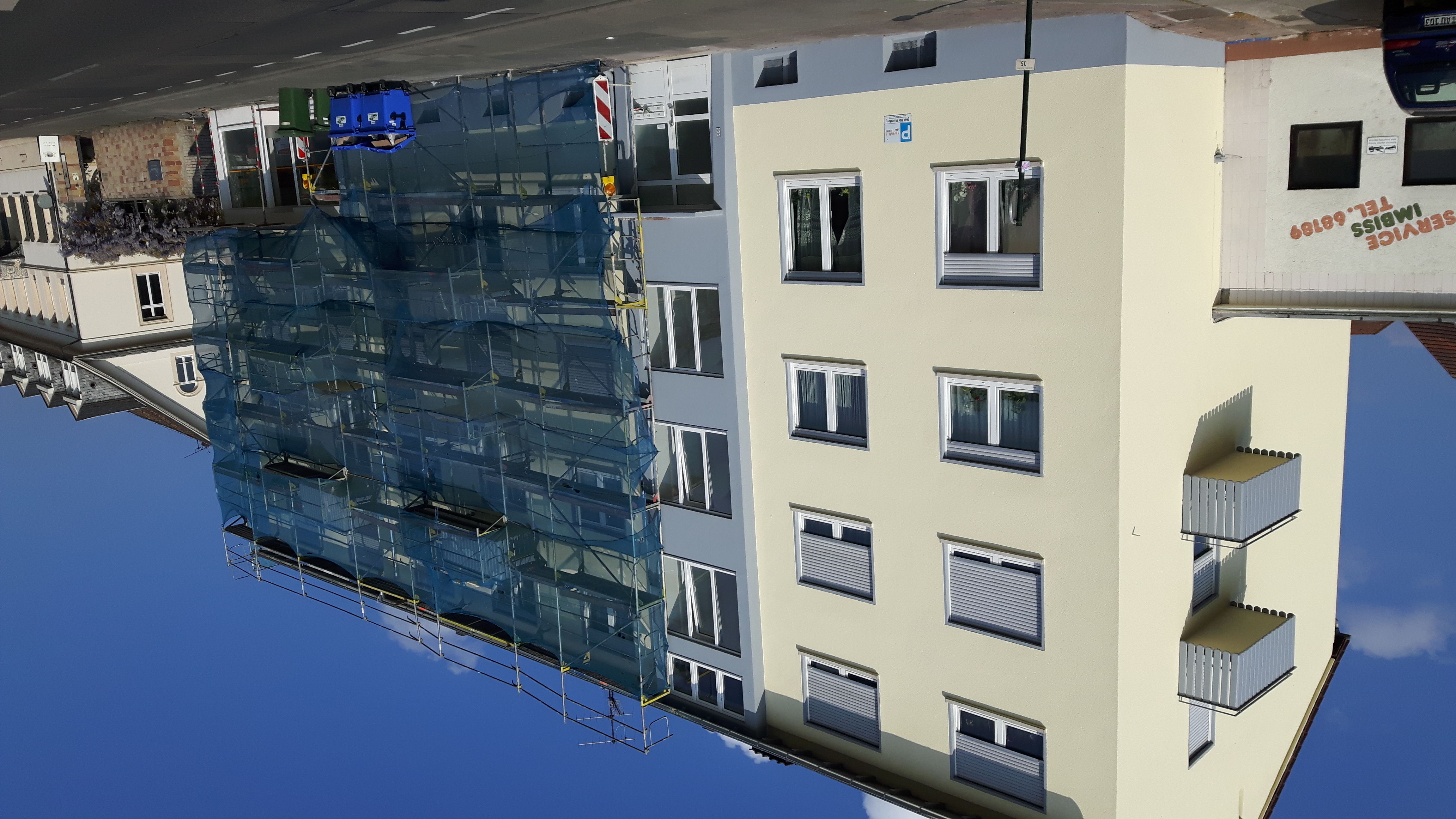 Budynek wielorodzinny w trakcie prac elewacyjnych, widoczne rusztowania z siatką ochronną na jednej części budynku, druga część z oknami i małymi balkonami, jasne kolory elewacji.