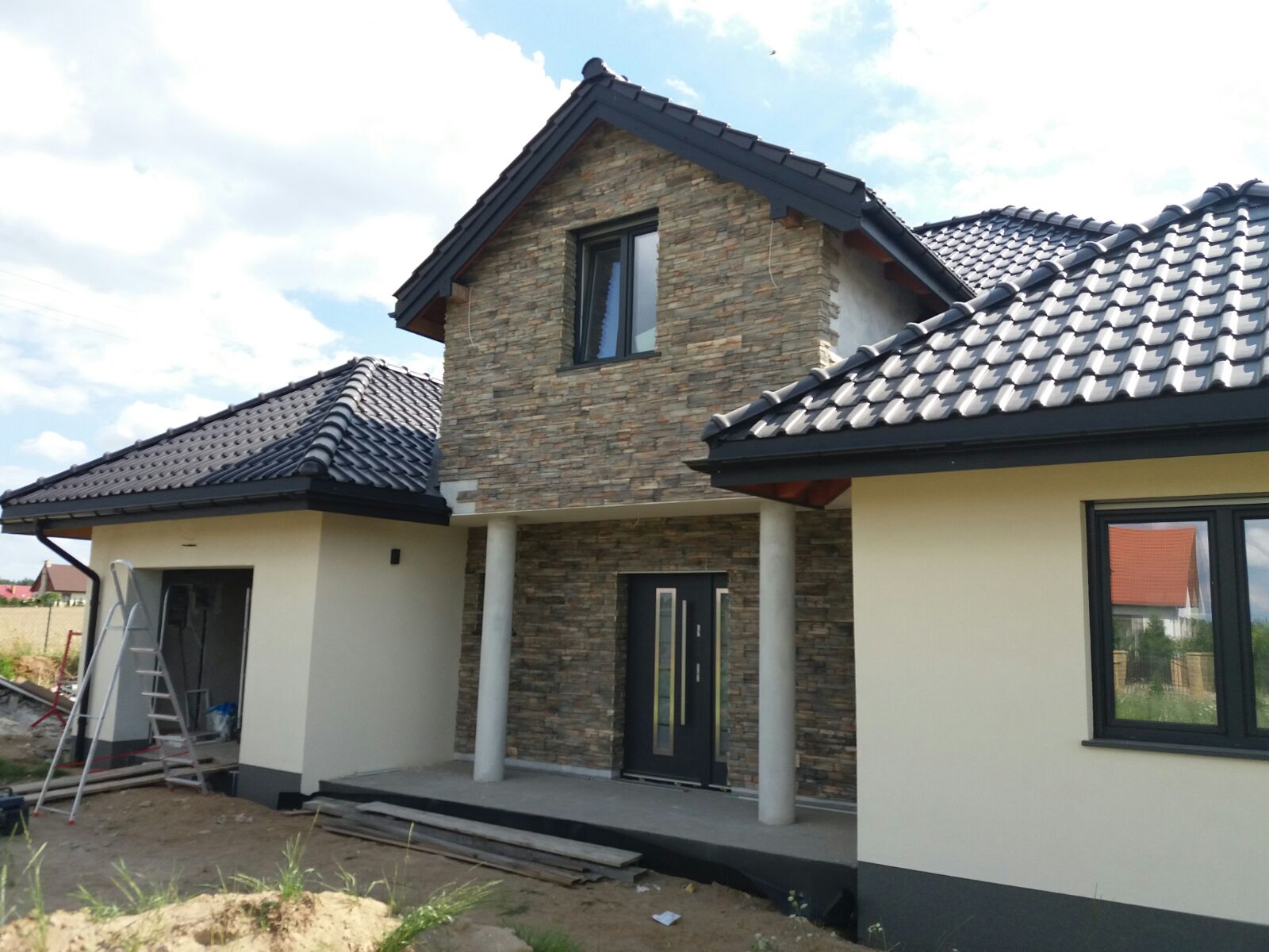 Nowoczesny dom jednorodzinny z elewacją wykończoną kamieniem dekoracyjnym, czarne okna i dachówka, białe ściany, drabina oparta o garaż, teren wokół domu w trakcie zagospodarowywania.