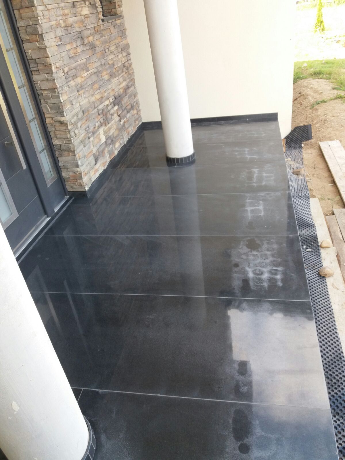 Wykończony taras z ciemnych, błyszczących płyt, z widocznym fragmentem elewacji z kamienia dekoracyjnego i kolumną podtrzymującą zadaszenie, na krawędzi tarasu widoczna siatka drenażowa.