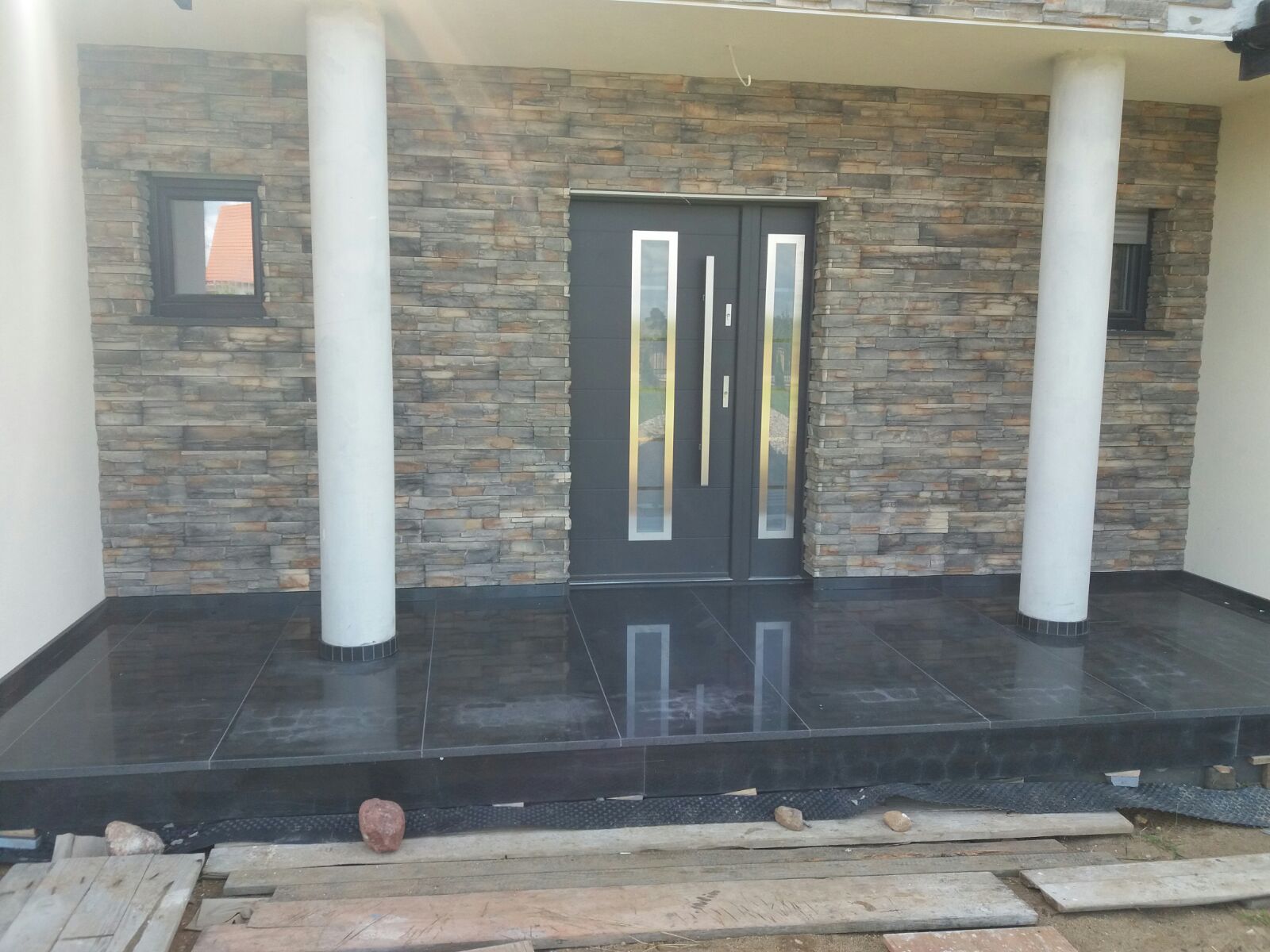 Elewacja budynku z kamienia dekoracyjnego, szara nowoczesna stolarka drzwiowa z przeszkleniami i czarna, błyszcząca posadzka z płytek na tarasie przed wejściem, widoczne drewniane elementy szalunku.