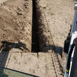 Wąski wykop w piaszczystej ziemi, widoczny z perspektywy kabiny koparki, z pracownikiem w oddali przy wykopie i zielenią w tle.