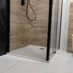 Nowoczesna kabina prysznicowa z czarnymi elementami, drewnopodobnymi płytkami na ścianach i białymi marmurkowymi płytkami na podłodze, widoczne baterie i deszczownica w kolorze czarnym.