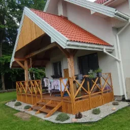 Nowoczesny dom z drewnianym tarasem i zadaszeniem, otoczony zielonym trawnikiem. Widoczne białe rynny i czerwona dachówka. Na tarasie suszarka z praniem.