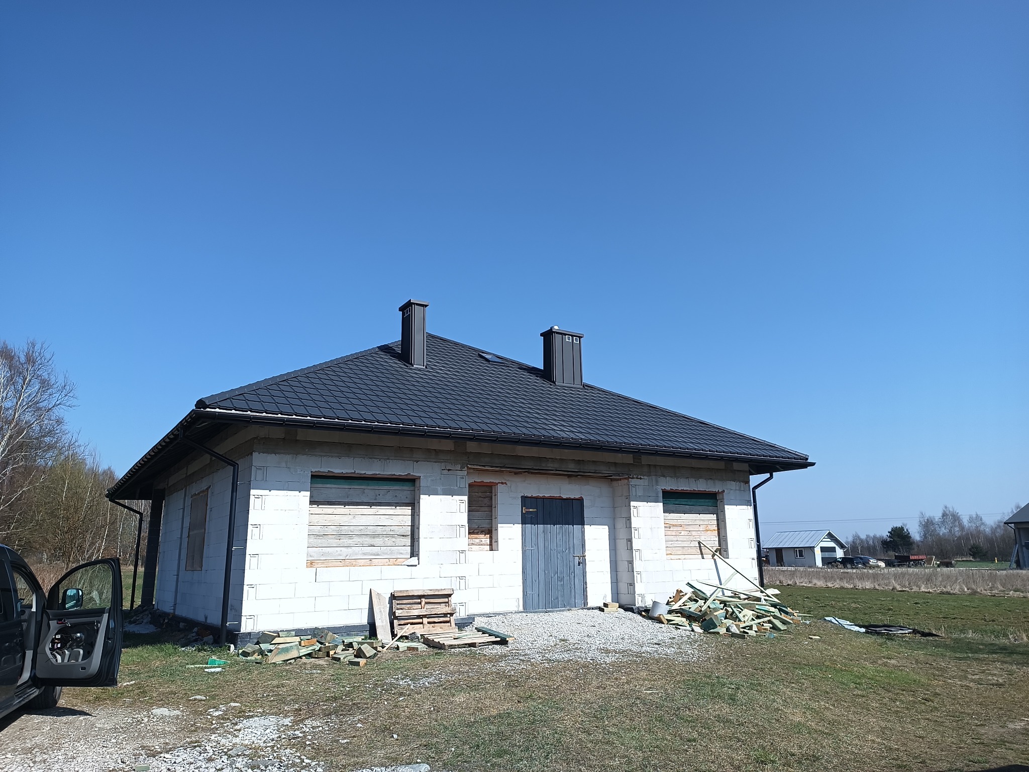 Niedokończony dom jednorodzinny z czarnym dachem i dwoma kominami, okna zabite deskami, otoczenie placu budowy z porozrzucanymi materiałami.