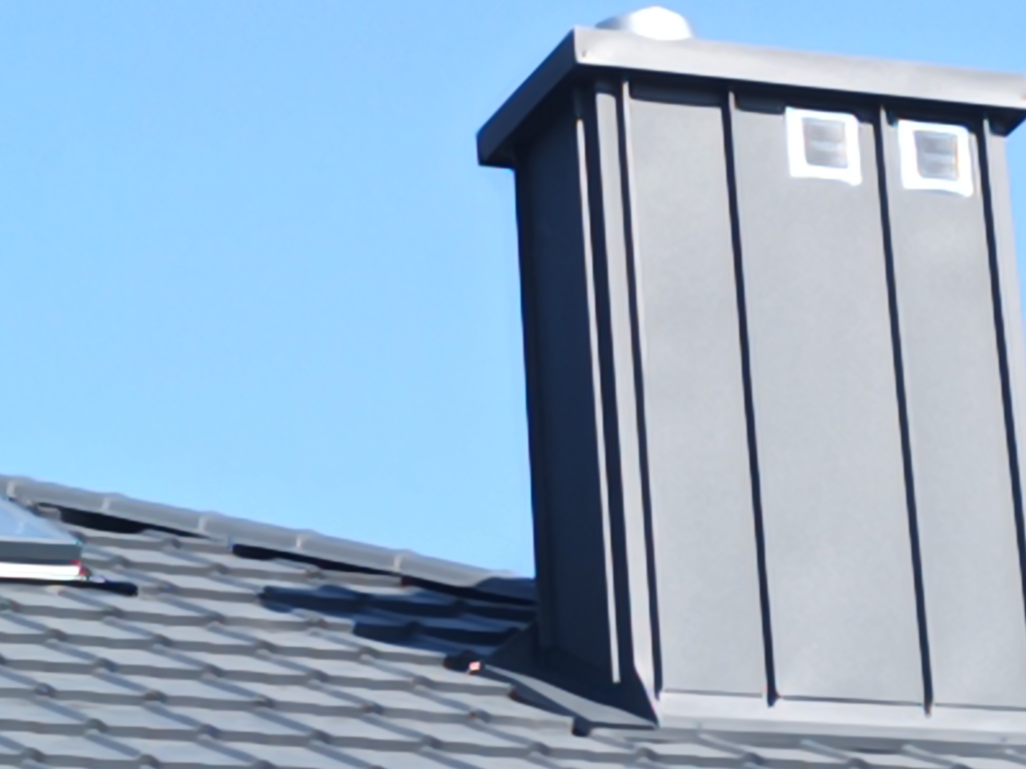 Szary komin z blachy z małymi okienkami wentylacyjnymi, wystający ponad dach pokryty szarą dachówką na tle błękitnego nieba.