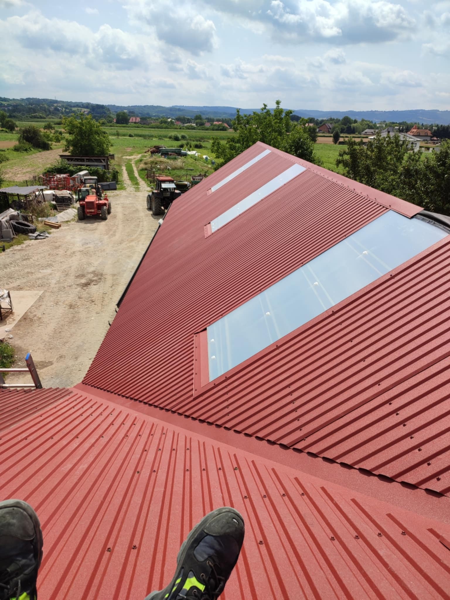 Czerwony, blaszany dach z trzema świetlikami w wiejskim otoczeniu, widok z góry, stopy w butach roboczych w dolnej części kadru.
