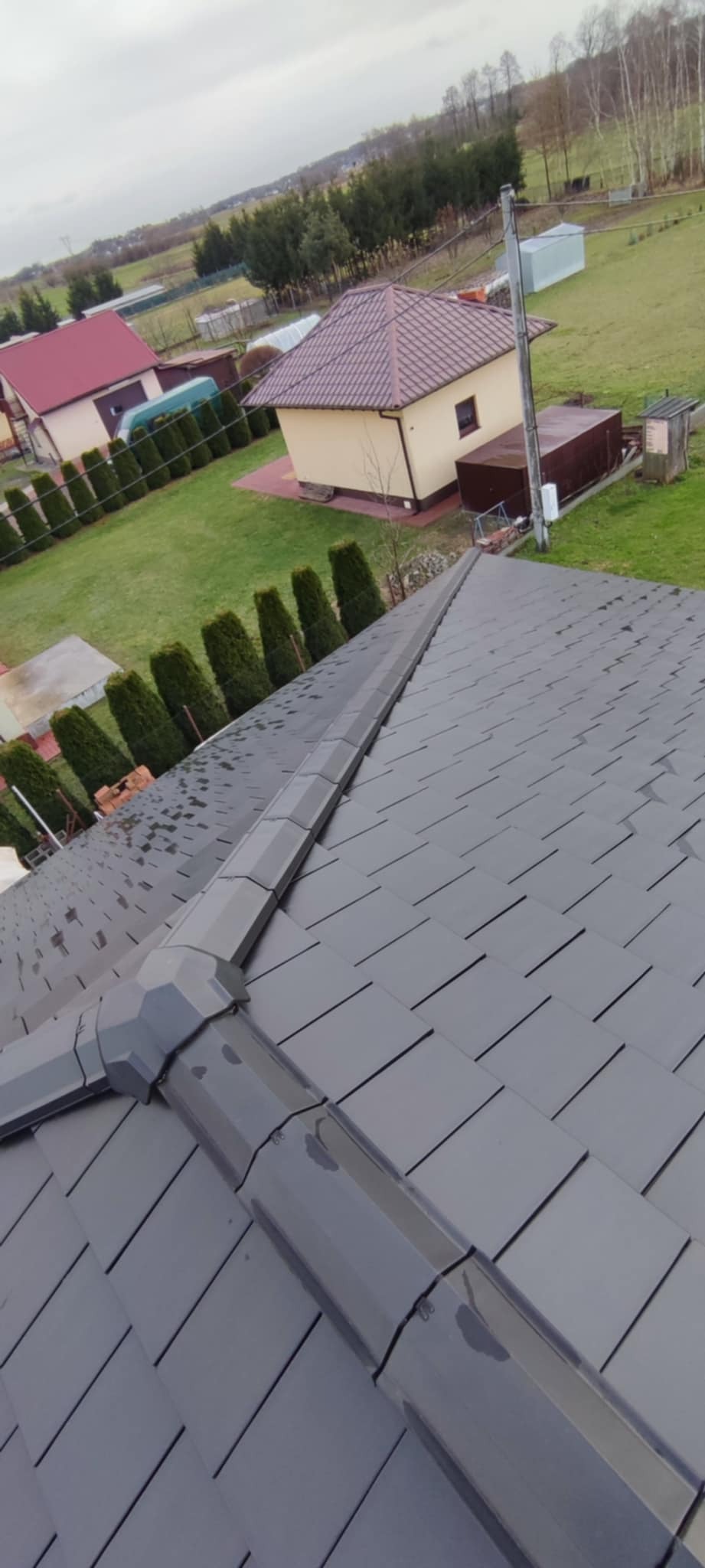 Nowy, grafitowy dach z dachówki bitumicznej z systemem rynnowym, widok z góry na tle zabudowań i zieleni w pochmurny dzień.