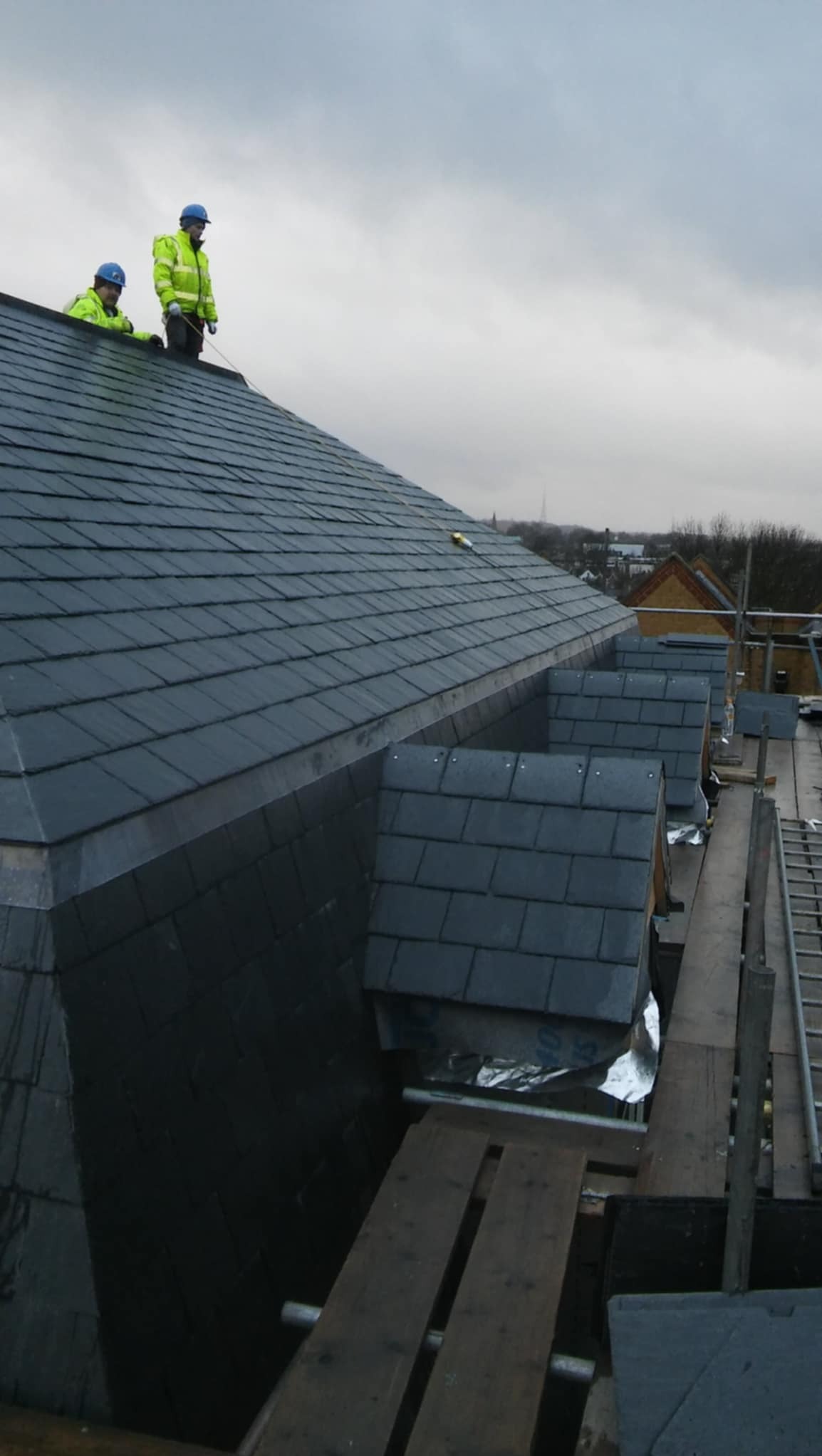 Dwóch dekarzy w odblaskowych kurtkach i kaskach pracuje na dachu pokrytym nową, ciemną dachówką łupkową, widoczne fragmenty rusztowania i lukarny.