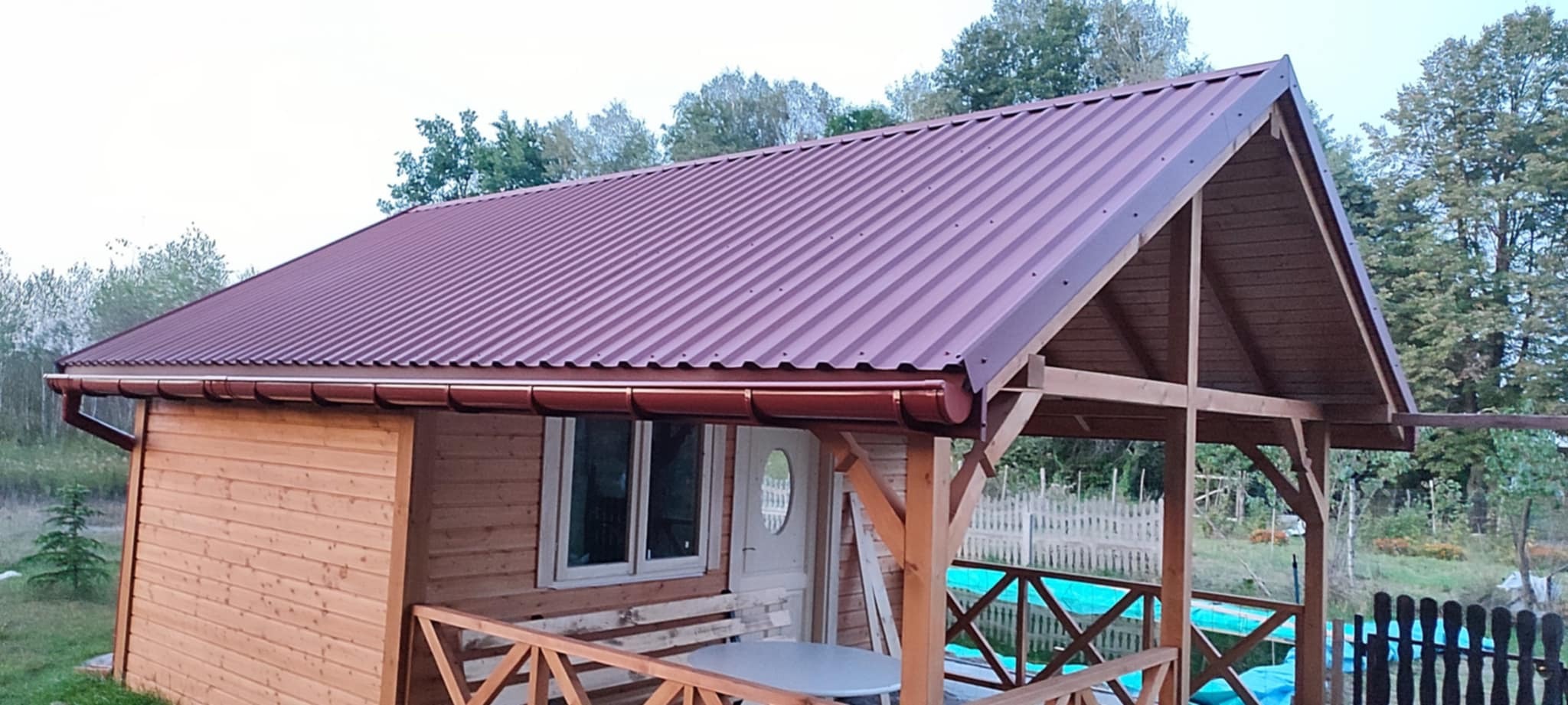 Nowy, bordowy dach z blachy trapezowej na drewnianym domku letniskowym z brązowymi rynnami, widok z zewnątrz na tle zieleni.