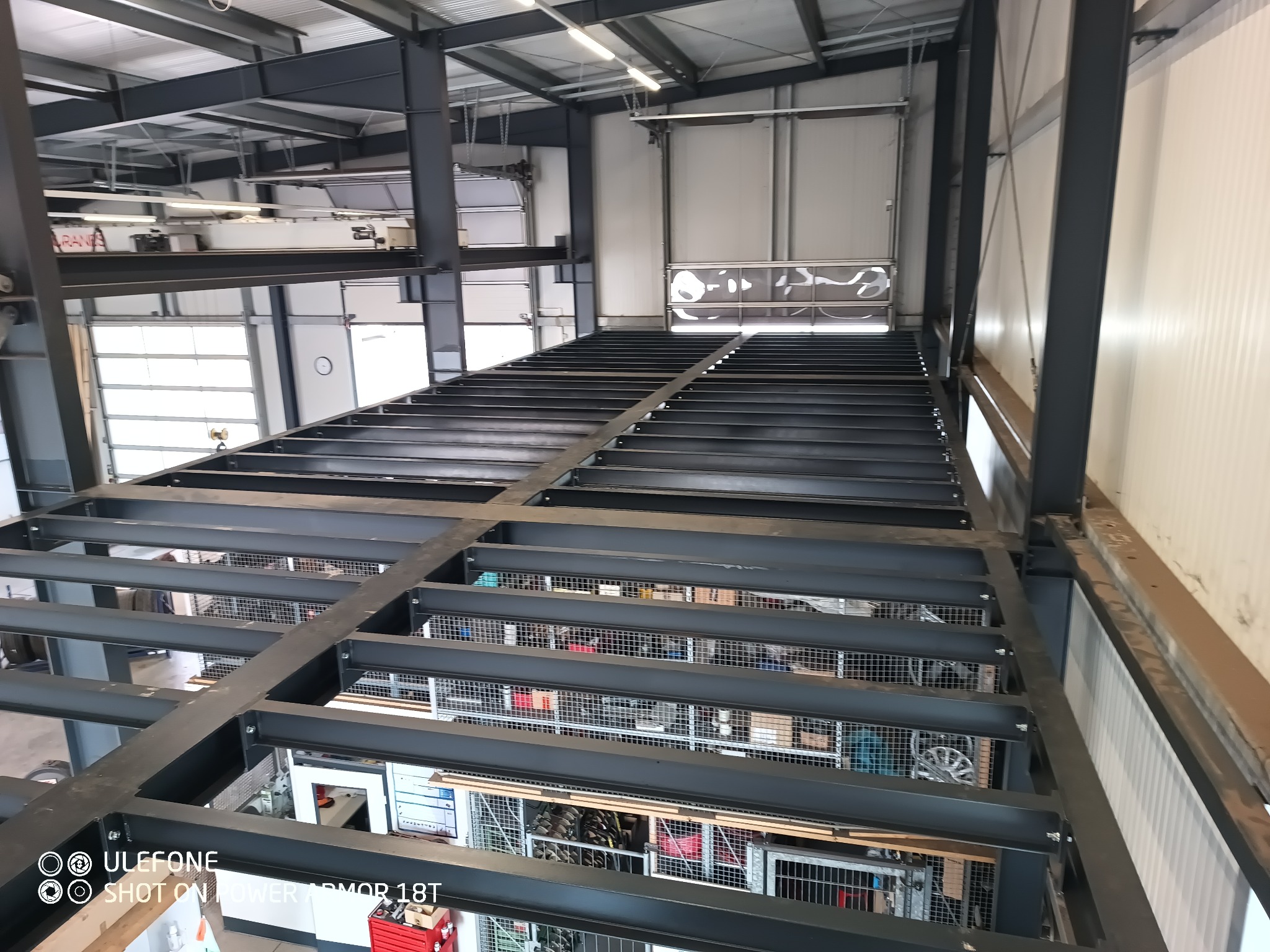 Konstrukcja stalowa antresoli w hali produkcyjnej, widok z góry na surowy szkielet podłogi z kratownicami, w tle białe ściany i bramy garażowe.