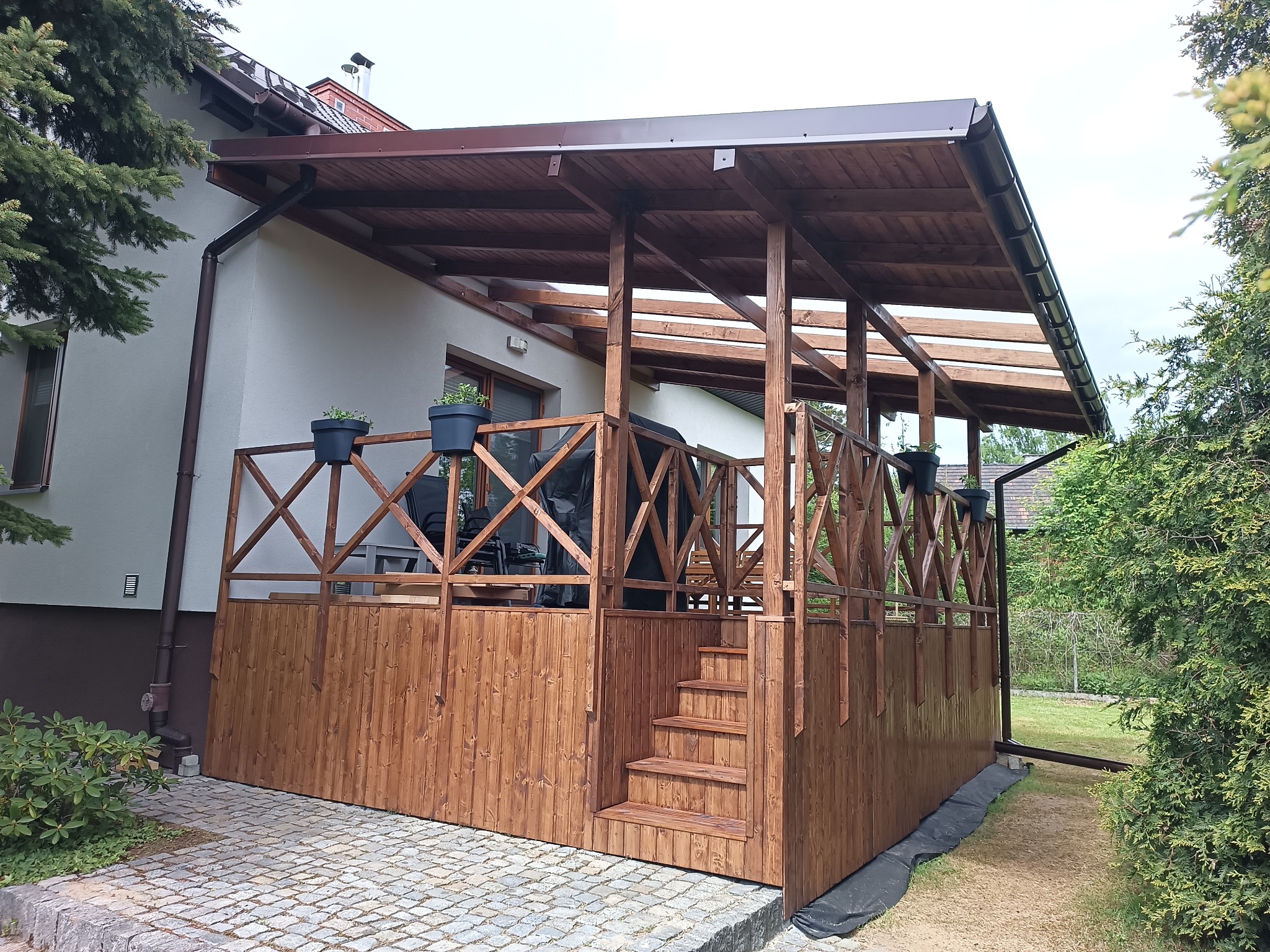 Zadaszony taras drewniany z barierką krzyżową i schodami, obok domu z brązową rynną. Na tarasie donice z kwiatami. Widok z poziomu gruntu, kamienna nawierzchnia.