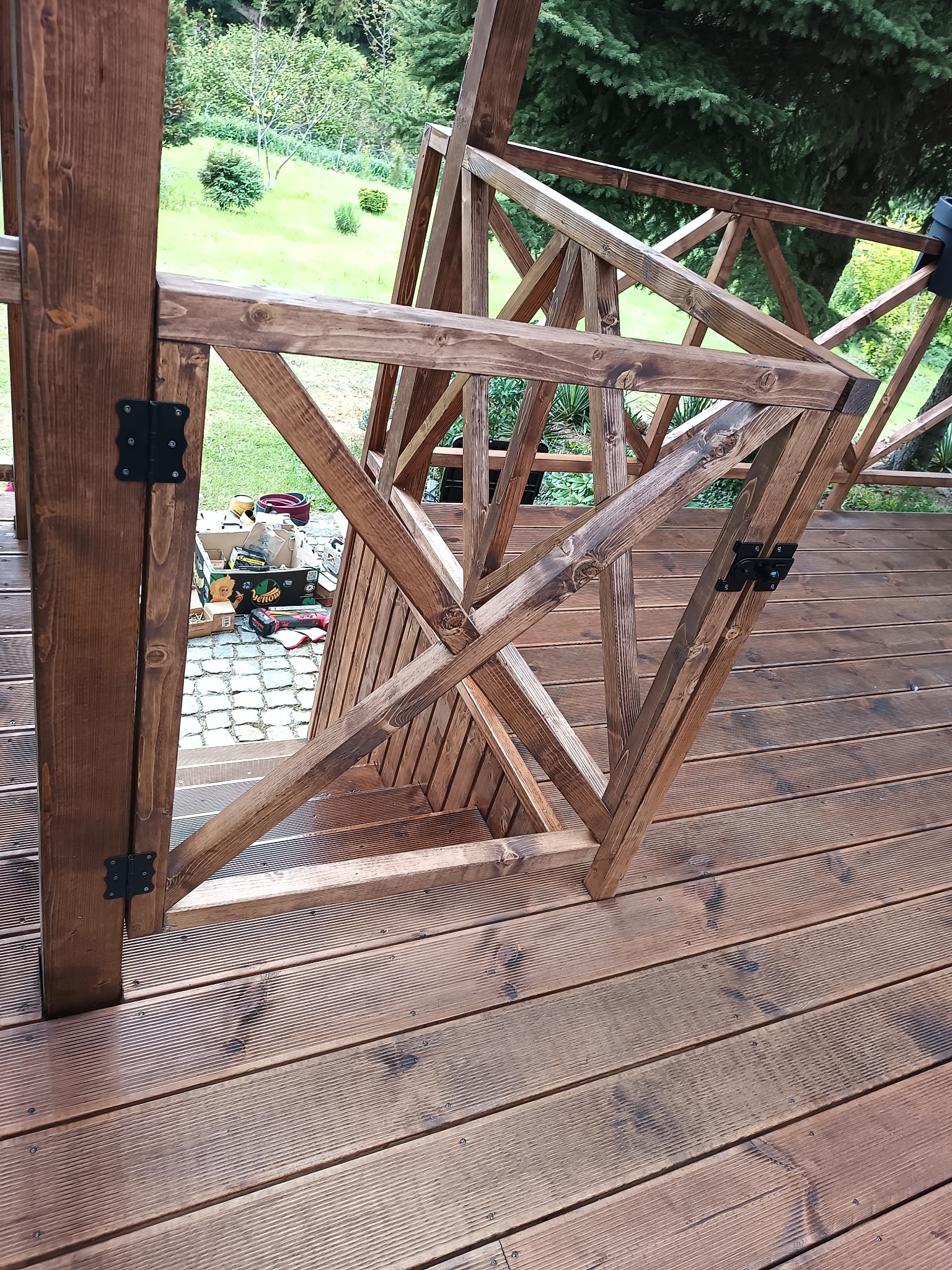 Drewniana konstrukcja tarasu w trakcie budowy, widoczne elementy balustrady i podłogi z desek ryflowanych. W tle zieleń ogrodu i narzędzia pracy.