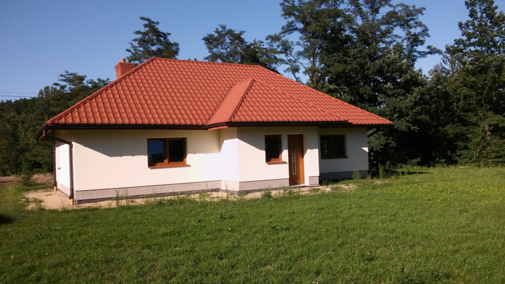 Dom jednorodzinny z czerwoną dachówką ceramiczną, widoczny z poziomu trawnika w słoneczny dzień. Okna i drzwi z brązowymi ramami.