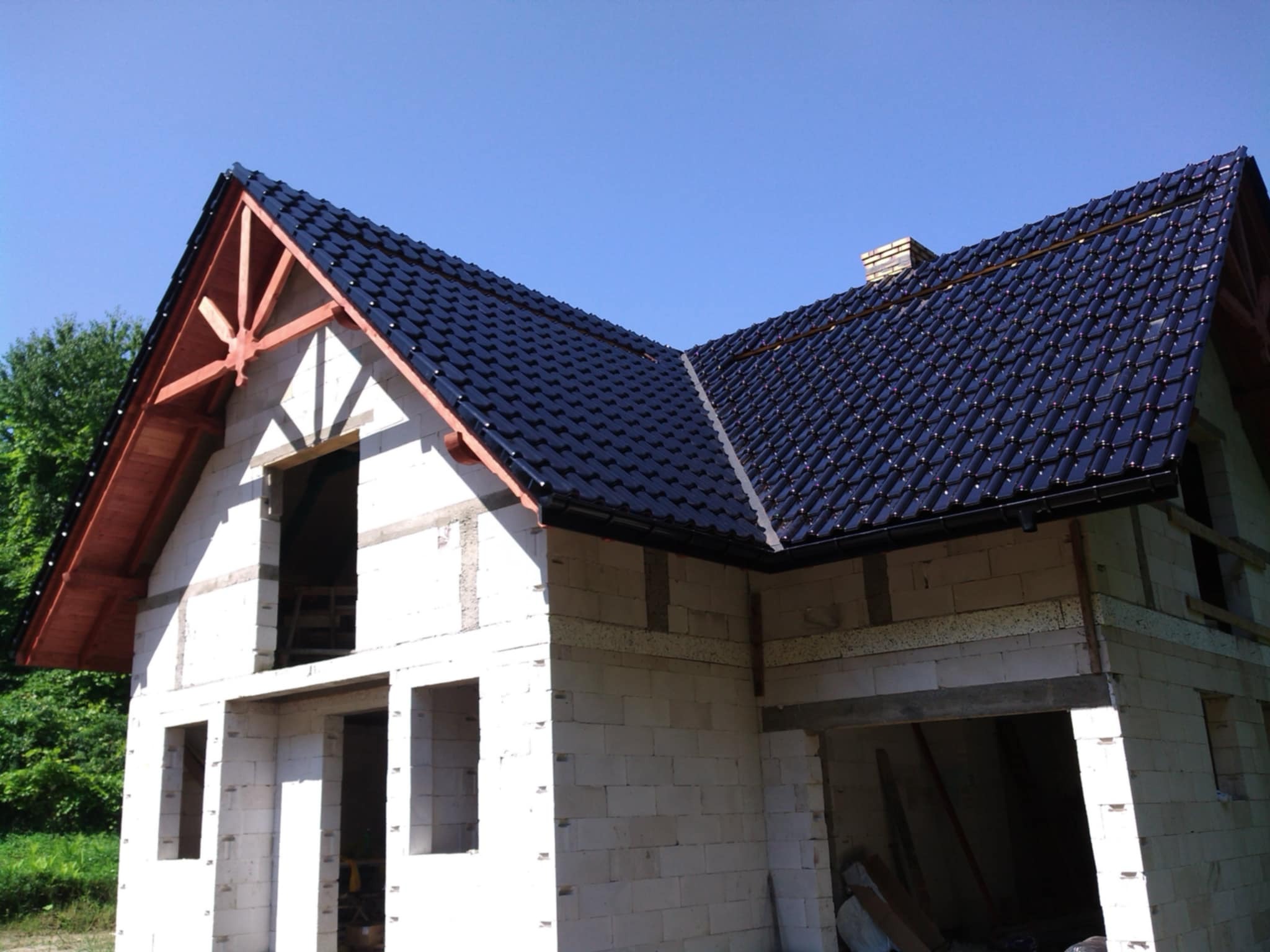 Niedokończony dom z czarnym dachem z dachówki ceramicznej w słoneczny dzień. Widoczne drewniane elementy konstrukcji dachu.