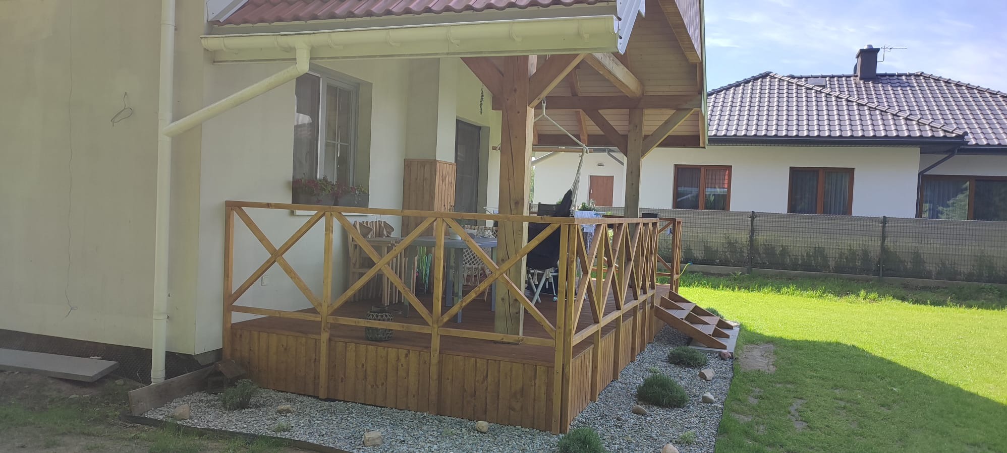Zadaszony taras drewniany z balustradą krzyżową, schodkami i obrzeżem z kamyków. Na tarasie stolik i krzesła. W tle dom z dachem z dachówki.