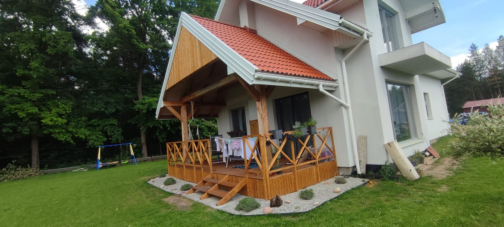 Nowoczesny dom z drewnianym tarasem i zadaszeniem, otoczony zielonym trawnikiem. Widoczne białe rynny i czerwona dachówka. Na tarasie suszarka z praniem.